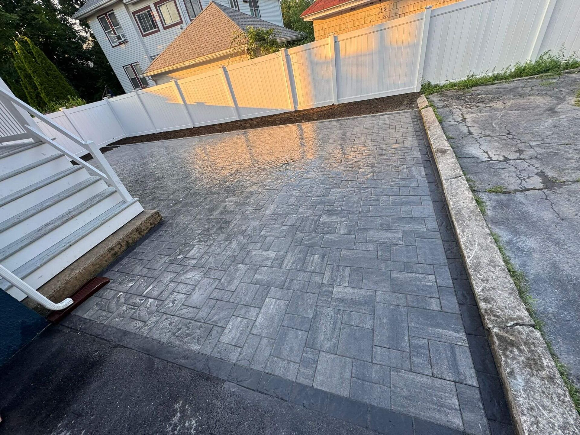 Brick-paved driveway beside a white fence and house steps, with a paved path and grassy slope nearby