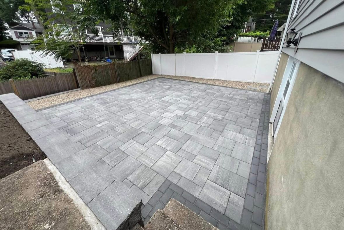 Backyard with newly paved gray stone patio beside a house and white fence