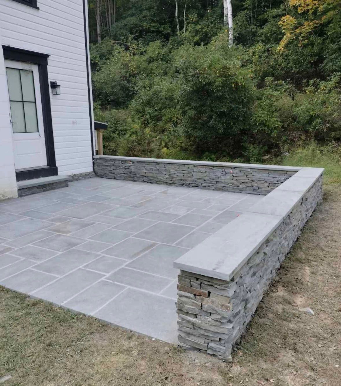 Stone patio and low retaining wall beside a house, with a grassy yard and wooded background.