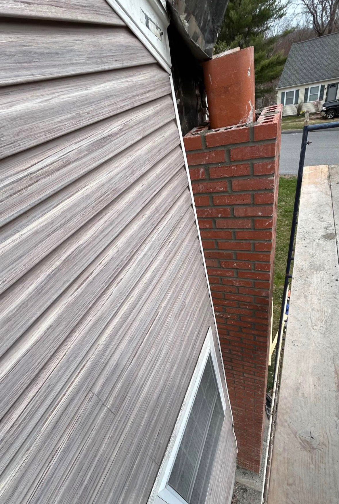 Narrow side view of a house exterior with tan siding, a brick wall, and a basement window well