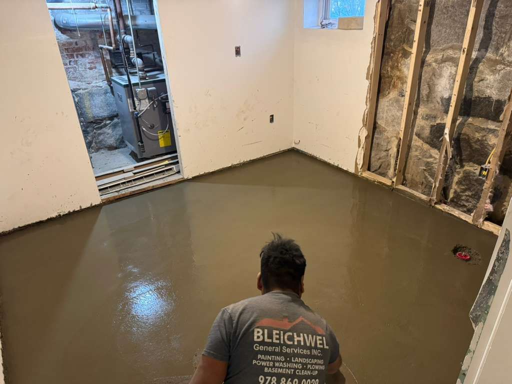 Worker smoothing wet concrete floor in an unfinished room with exposed walls and an open doorway