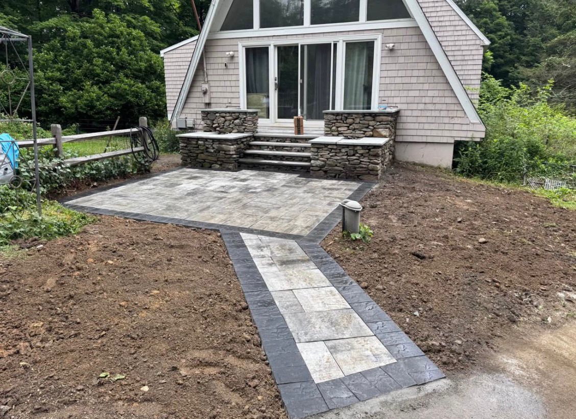 Stone patio and walkway leading to a small house with an A-frame roof, surrounded by bare soil and greenery