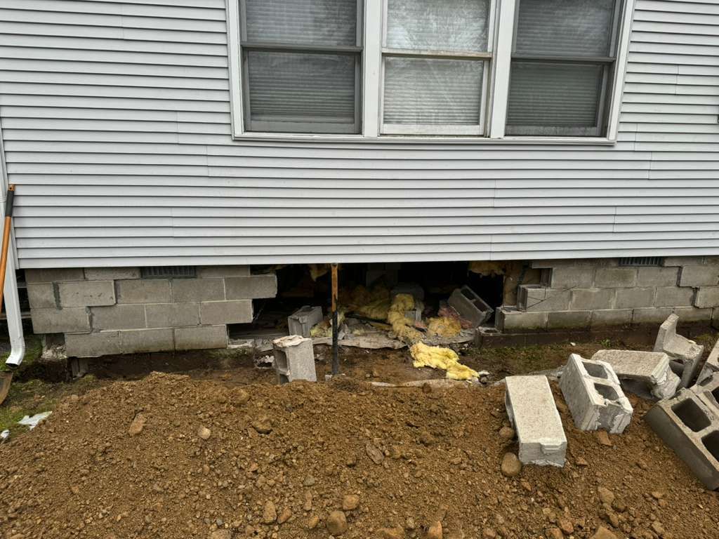 House foundation with missing cinder blocks, exposed insulation, and dirt piled beneath the siding