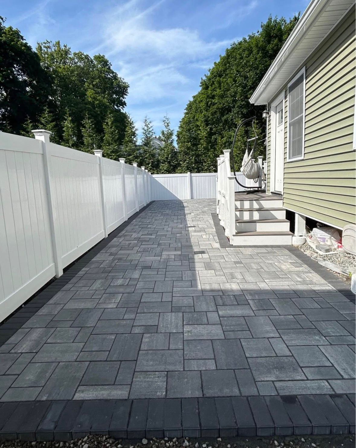 Paver patio and walkway beside a white vinyl fence and house, leading to a backyard seating area