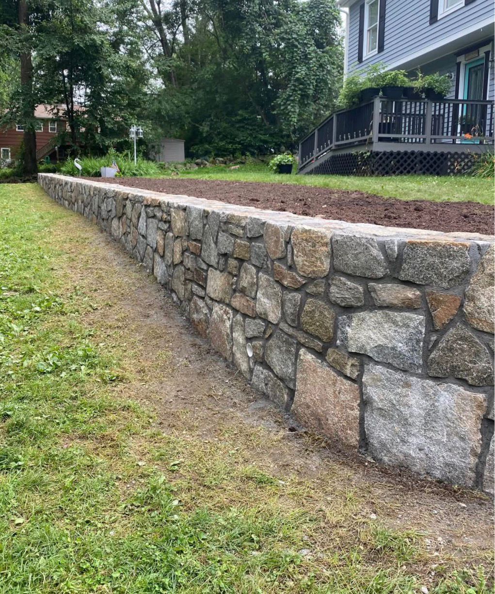 Stone retaining wall beside a grassy yard and house, with a sloped dirt bed above it.