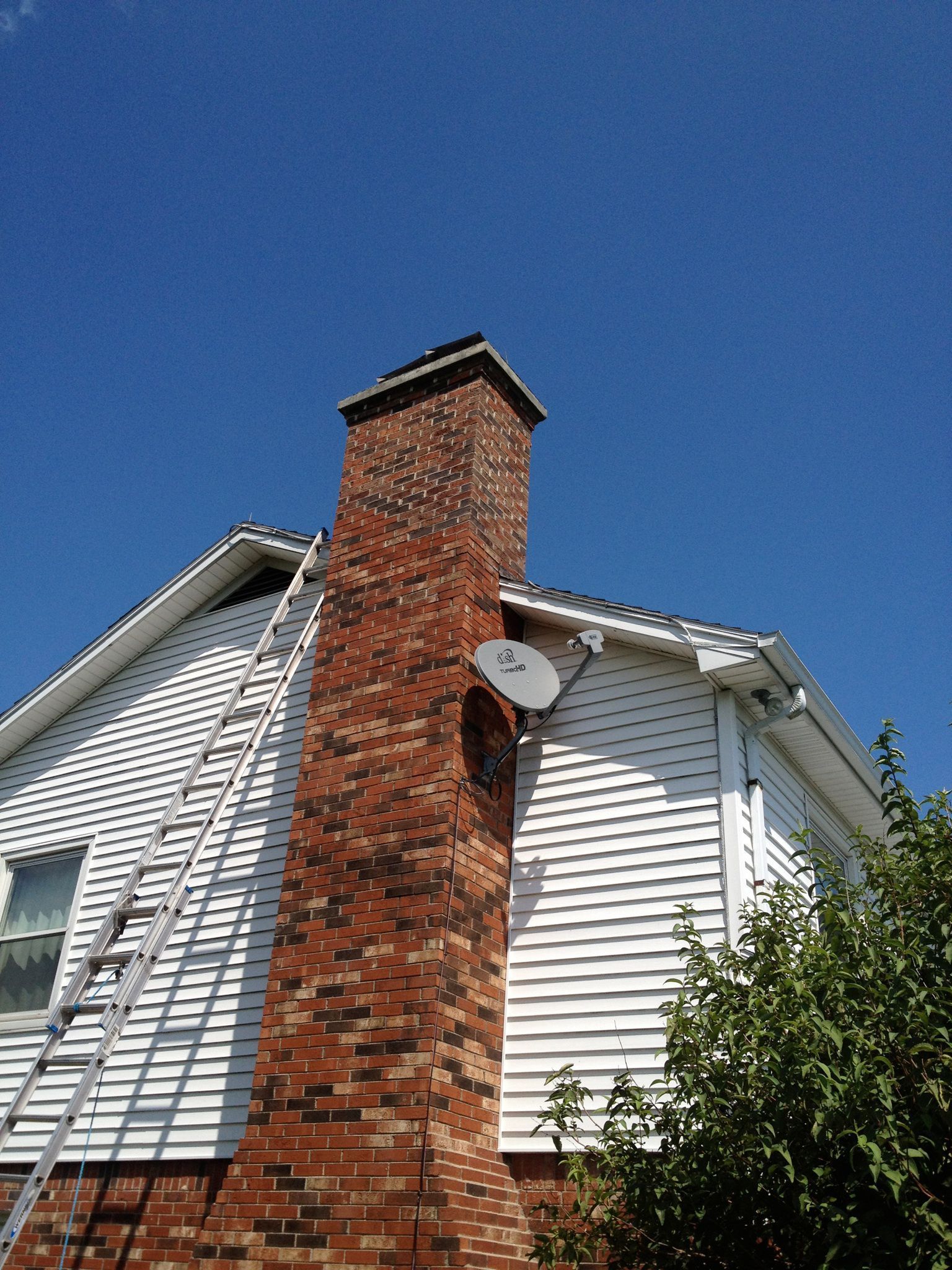 Brick chimney with a satellite dish on a white house against a clear blue sky