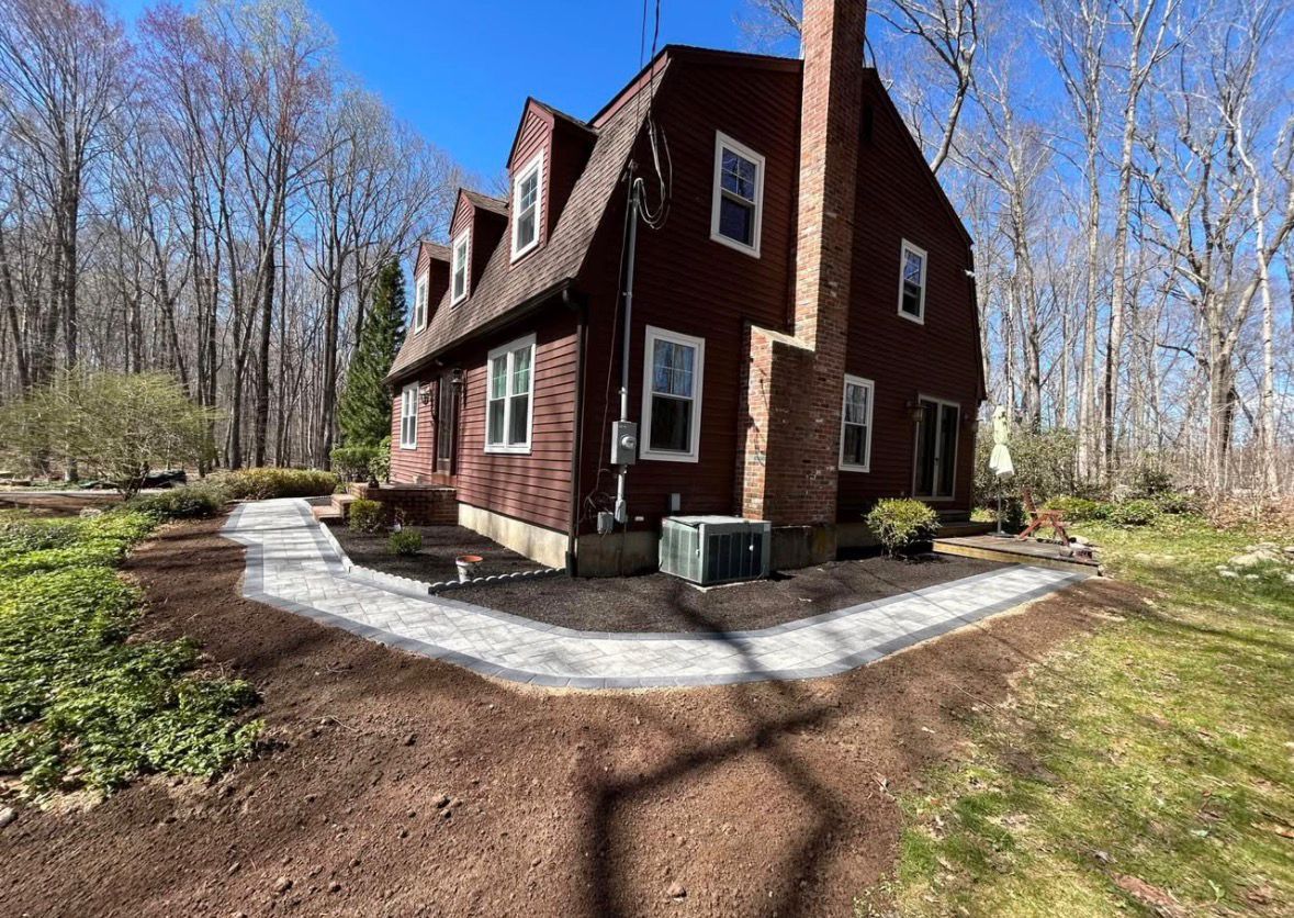 Red cabin-style house with white trim in a wooded yard, surrounded by a gravel path and spring trees