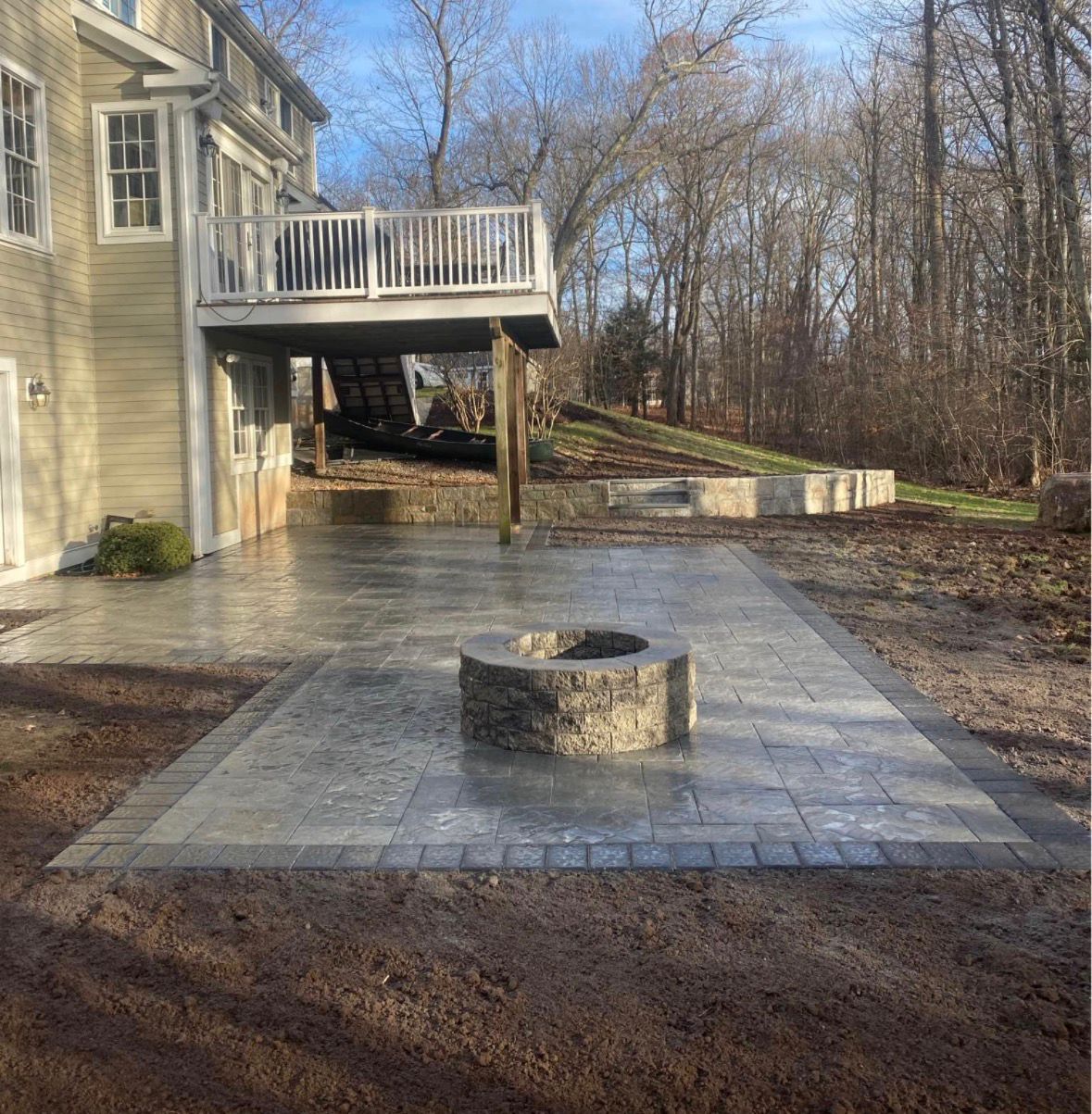 Concrete patio with circular fire pit beside a beige house and deck, bordered by woods.