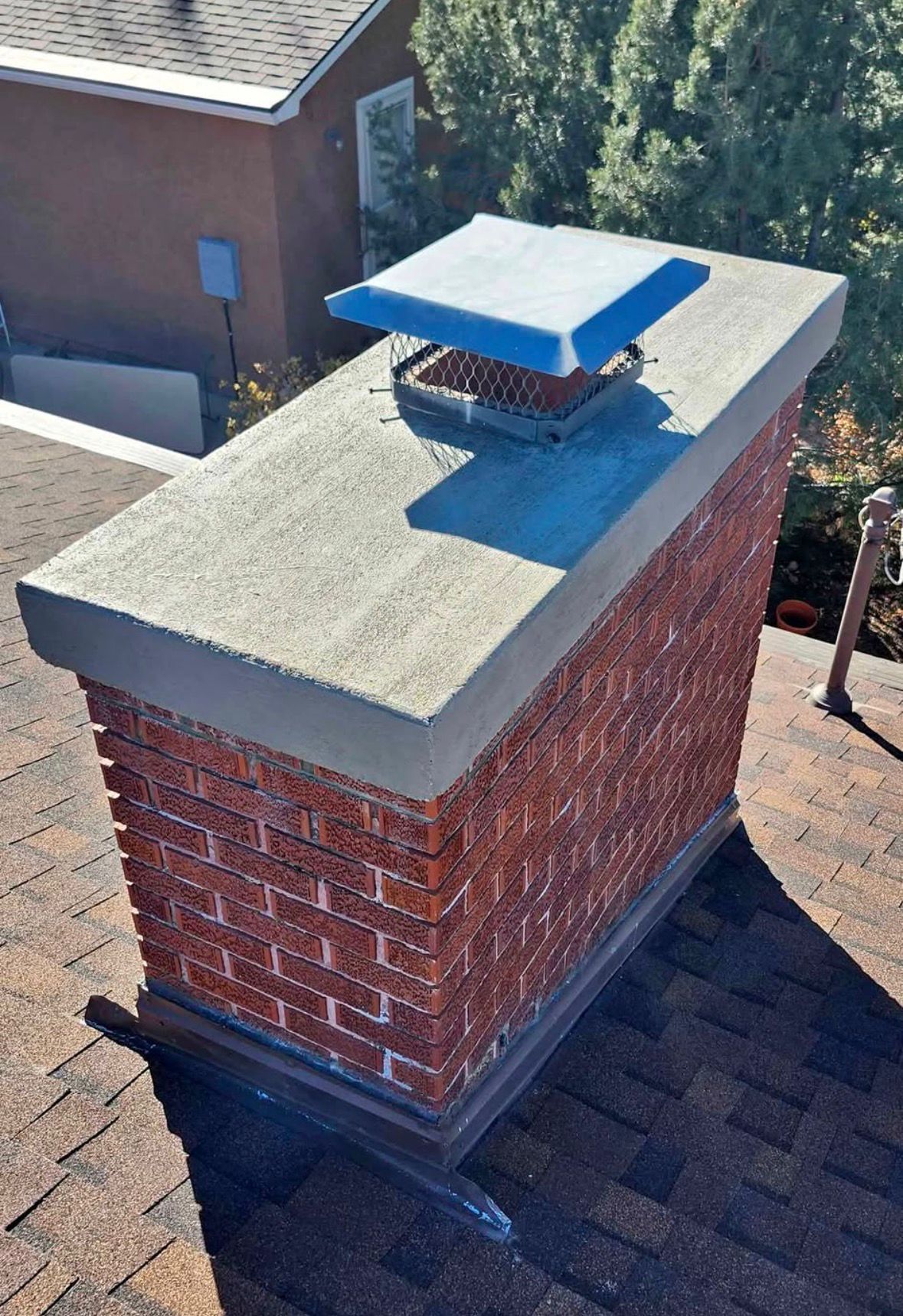 Brick rooftop chimney with a concrete cap and blue vent on a shingled roof.