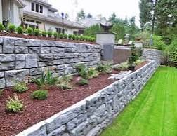 Stone retaining wall beside landscaped garden with mulch, shrubs, and green lawn.