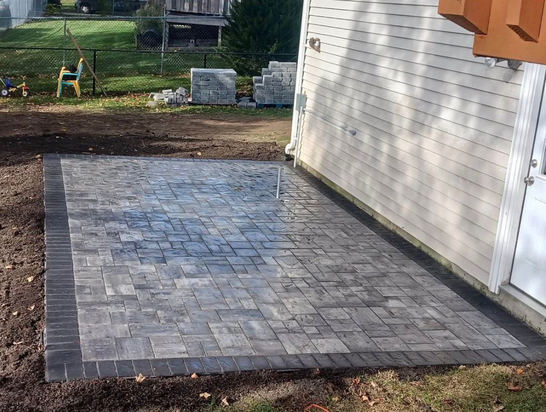 Paver patio beside a house with a dark border, set in a backyard with grass and trees