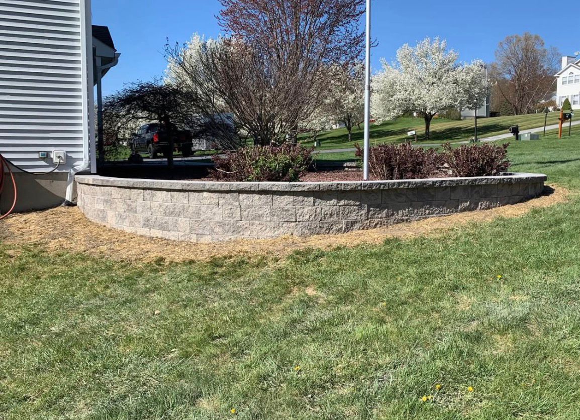 Stone retaining wall bordering a grassy yard with blooming trees beside a house