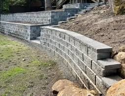 Curved gray retaining wall made of stacked blocks along a sloped yard with dirt and grass.