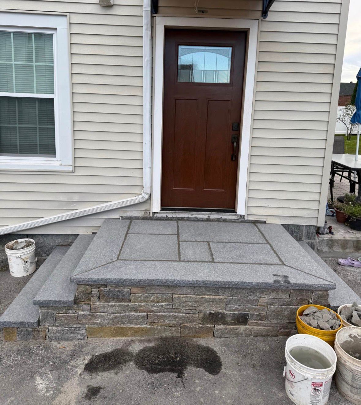 Front entry steps with gray pavers leading to a brown door on a beige-sided house