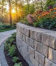 Curved stone retaining wall beside a landscaped garden path at sunset, with flowers and trees.
