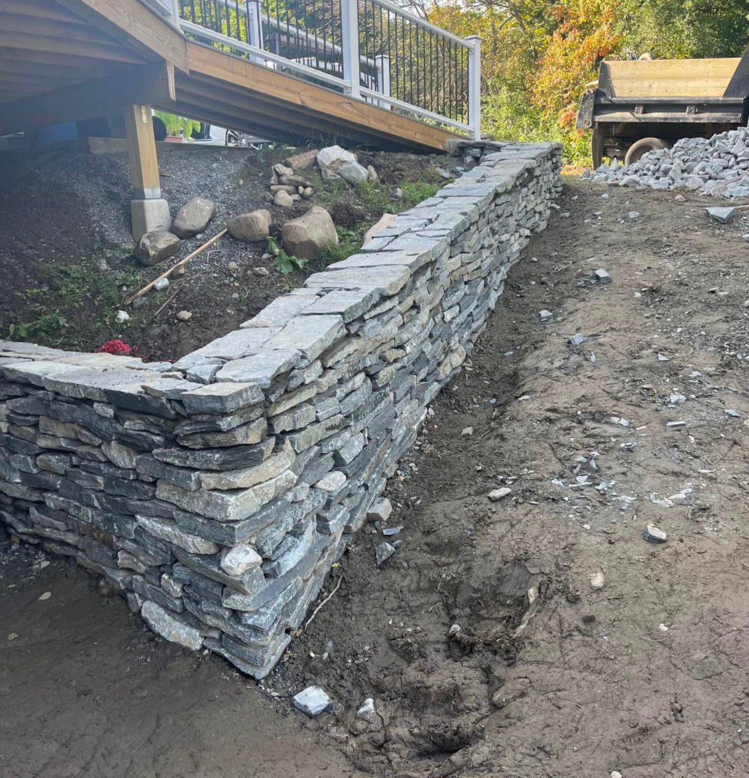 Stone retaining wall under construction beside a dirt slope and wooden deck.