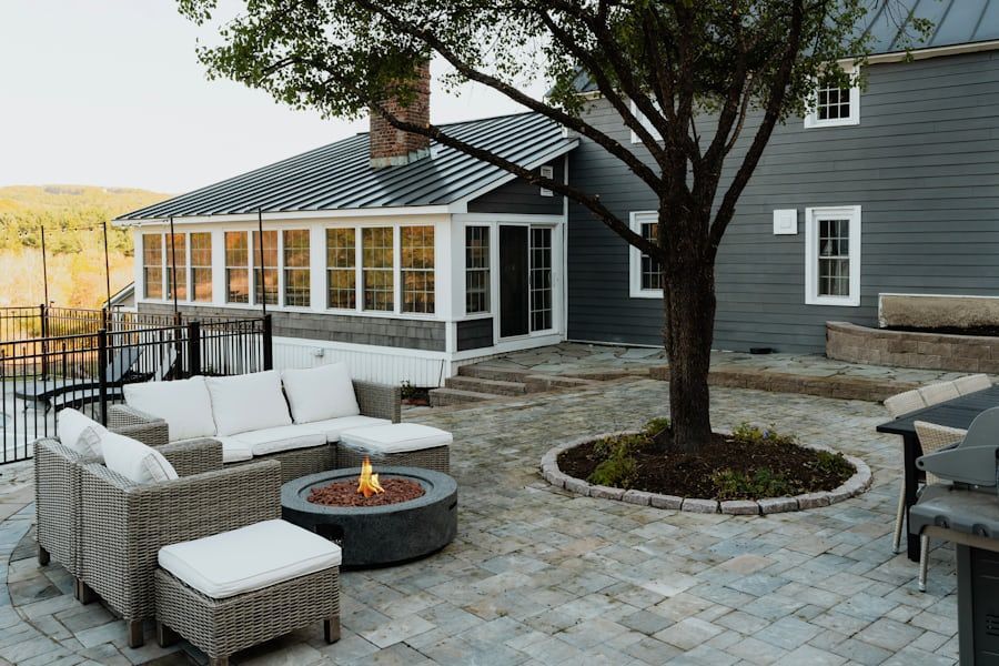 Patio with wicker seating, fire pit, and trees beside a gray house and sunroom