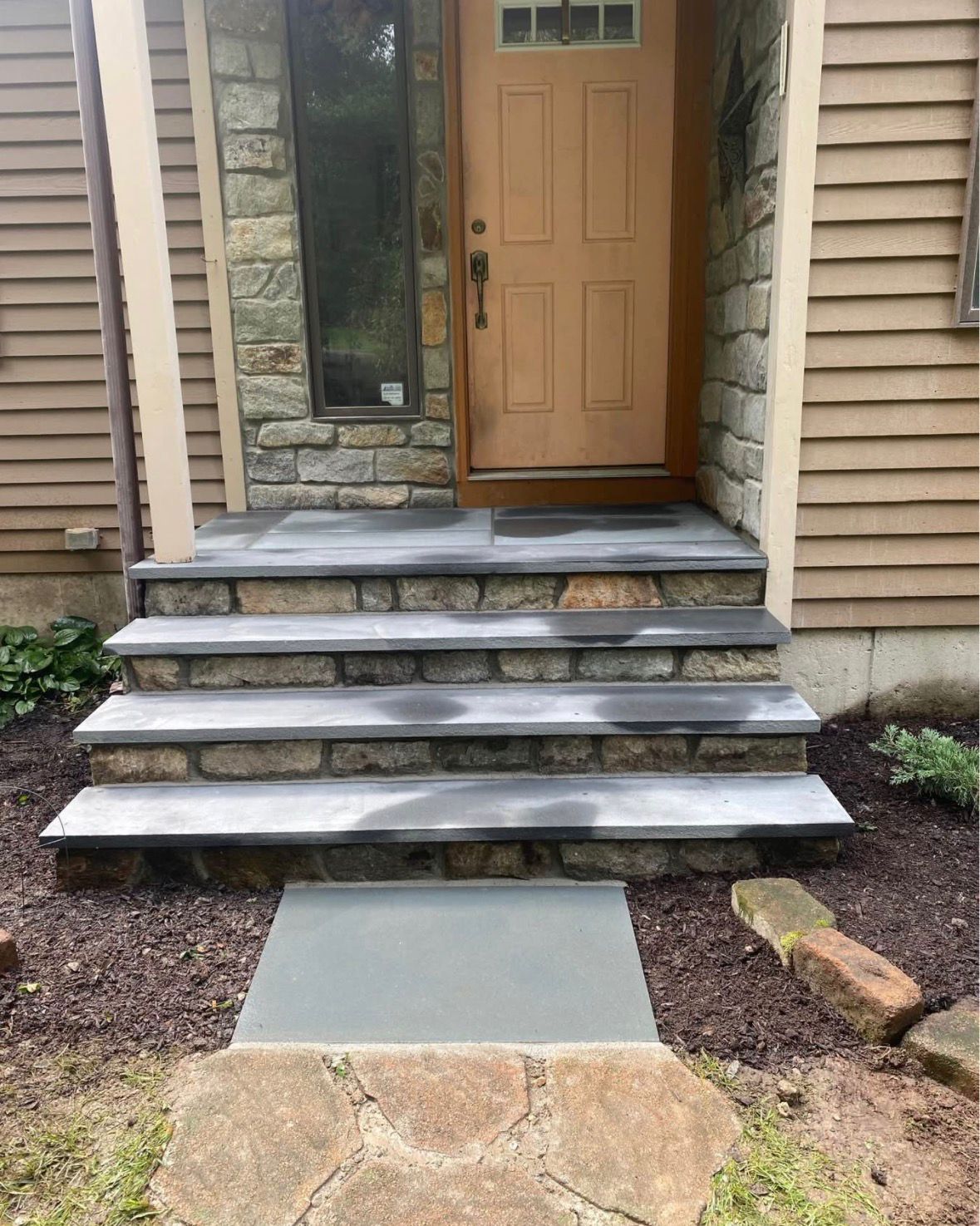 Front entrance with tan door, stone steps, and a concrete walkway leading to the porch.