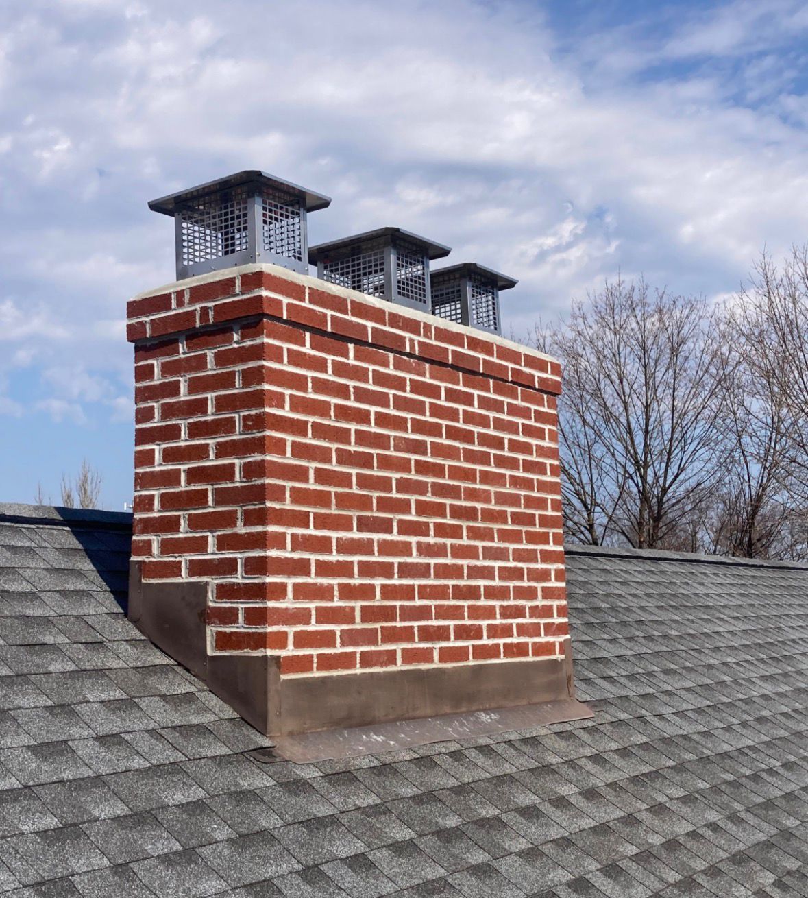 Brick chimney with three metal flue caps on a gray shingled roof under a cloudy sky