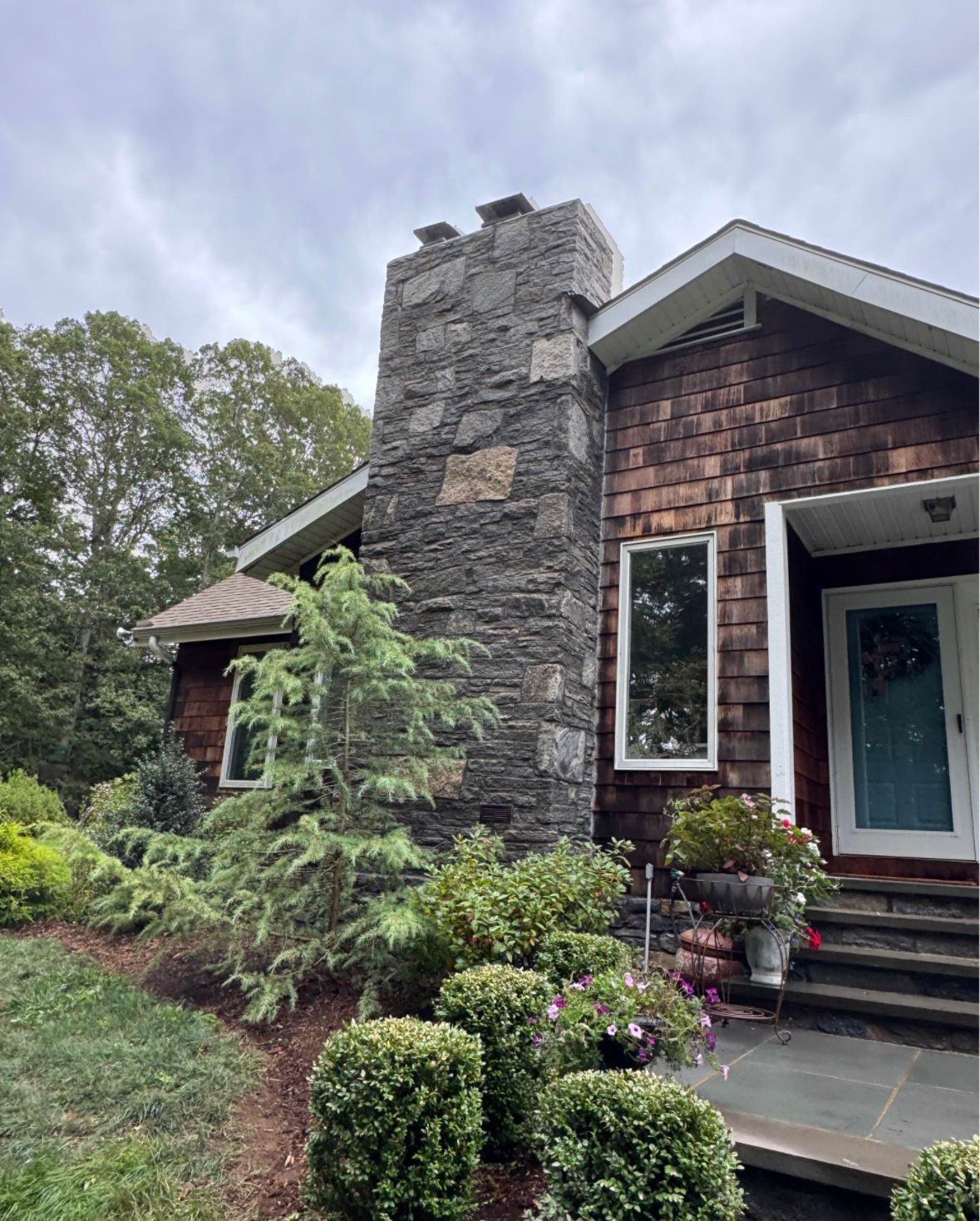 Brick house exterior with stone chimney, front steps, and lush shrubs under a cloudy sky