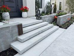Modern concrete steps leading to a front door, with potted red flowers and landscaped planters on both sides