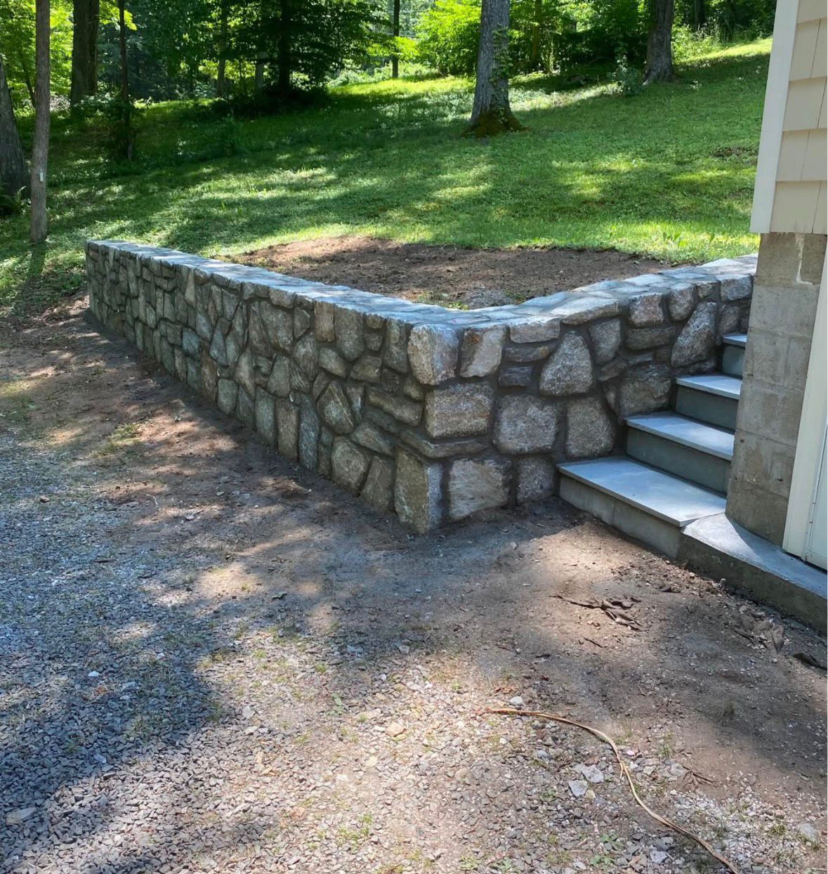 Stone retaining wall beside outdoor steps next to a house, with a grassy wooded slope behind it