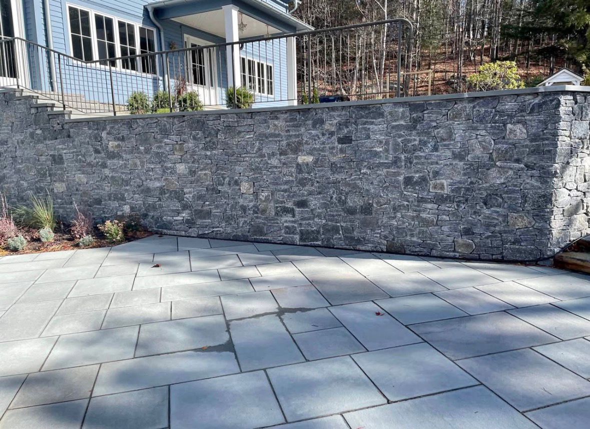Stone retaining wall beside a paved patio with a house porch and landscaping in the background
