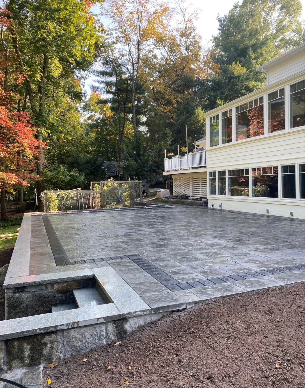 Large stone patio beside a white house, with a small recessed pool or fountain and autumn trees nearby