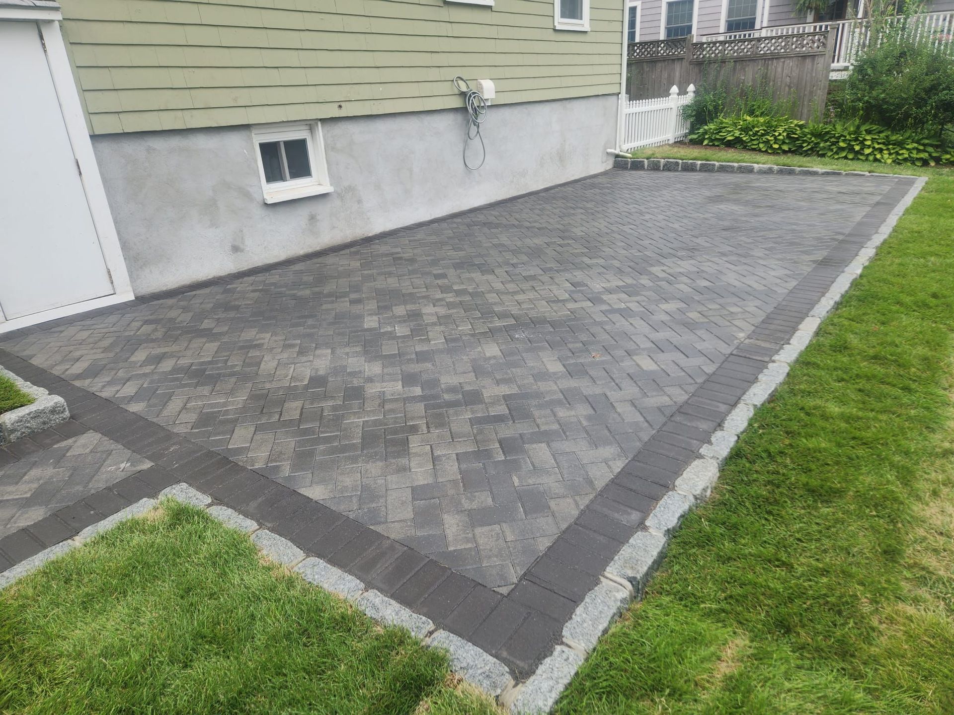 Gray paver patio bordered by dark bricks beside a house, with grass along the edge and a window nearby
