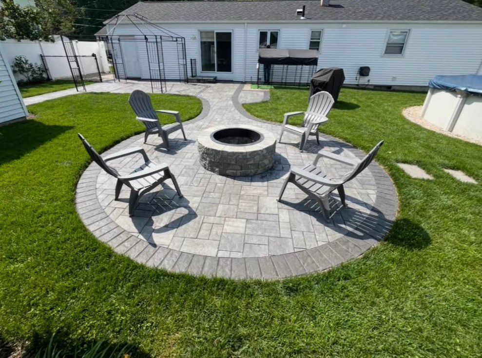 Patio with circular stone fire pit and four black chairs on a grass lawn beside a white house