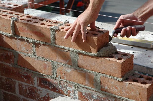 Hands laying red bricks on a partially built brick wall with mortar and tools nearby