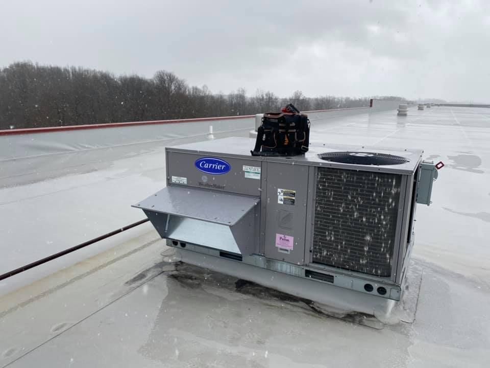 A Carrier rooftop HVAC unit sits on a wet, gray flat roof under an overcast, snowy sky.