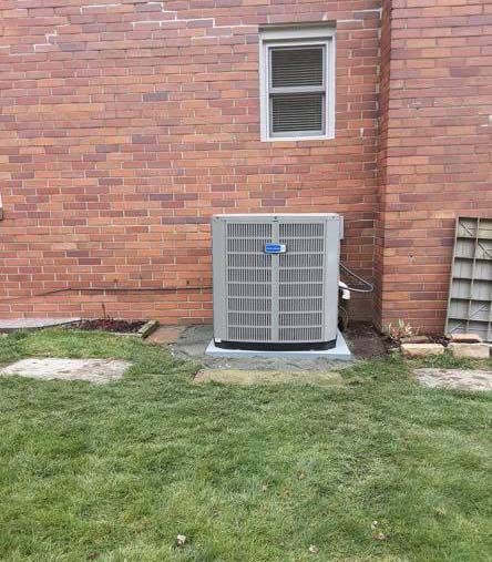 A grey HVAC unit sits on a concrete pad against a red brick wall with a small window above it in a grassy yard.