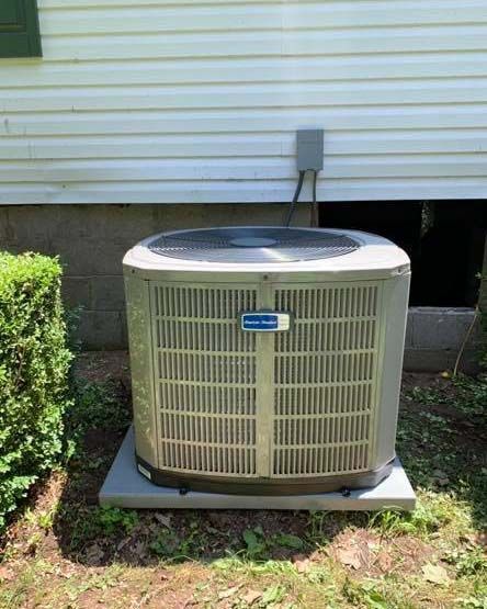 A light gray American Standard outdoor air conditioning unit sits on a concrete pad against a white-sided house.