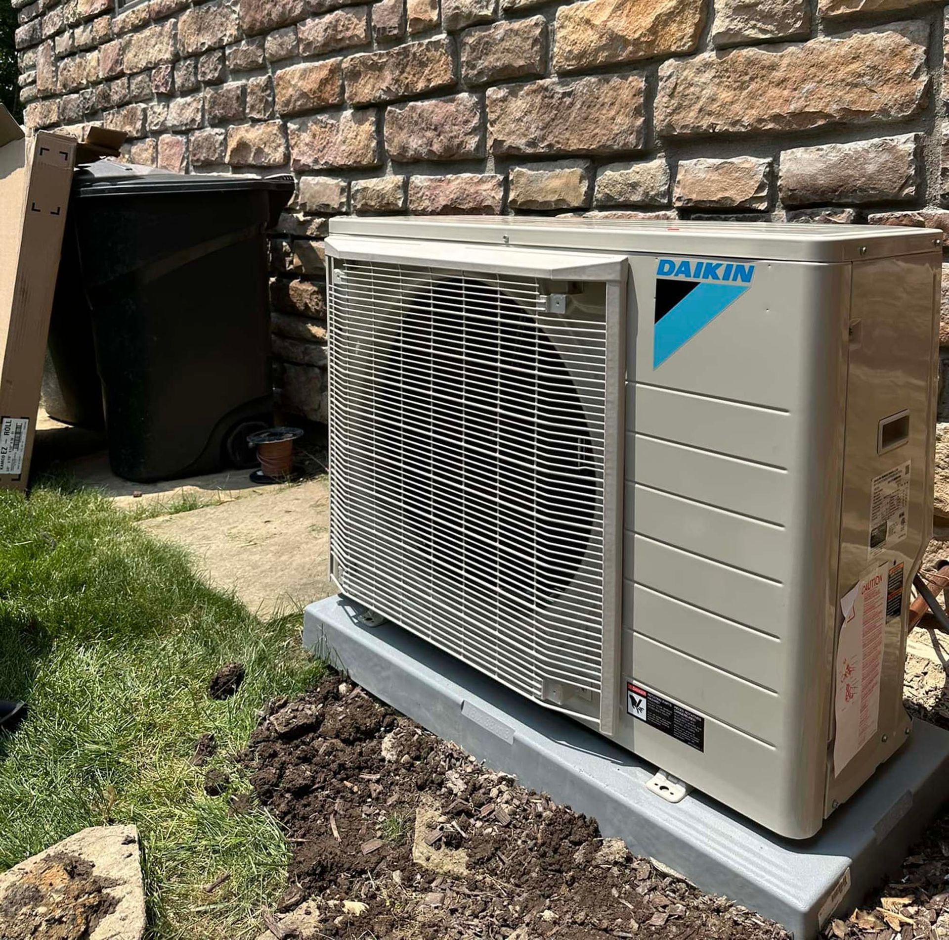 A Daikin outdoor HVAC unit sitting on a grey plastic base against a stone wall next to a black trash bin.