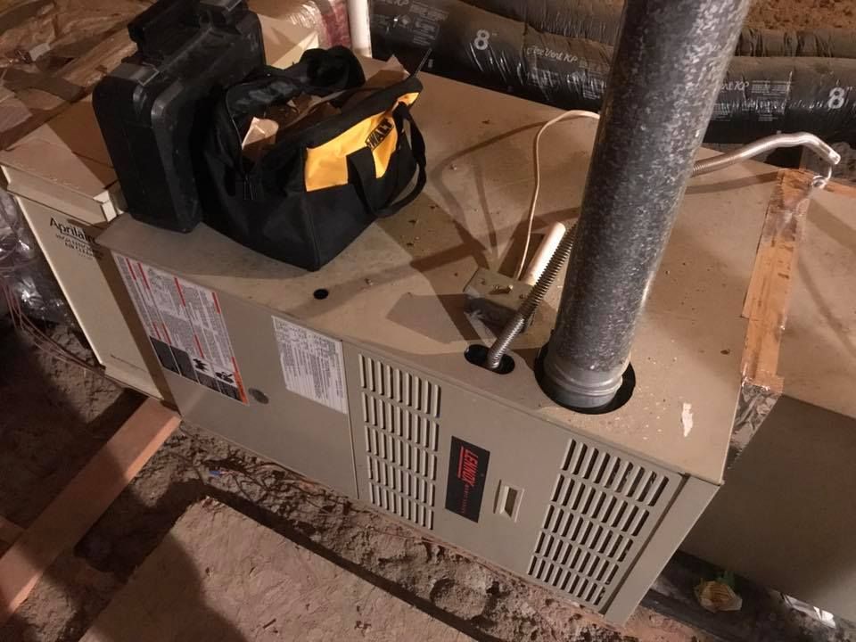 A furnace unit in an attic with a tool bag and a drill case resting on top, near a silver exhaust pipe.