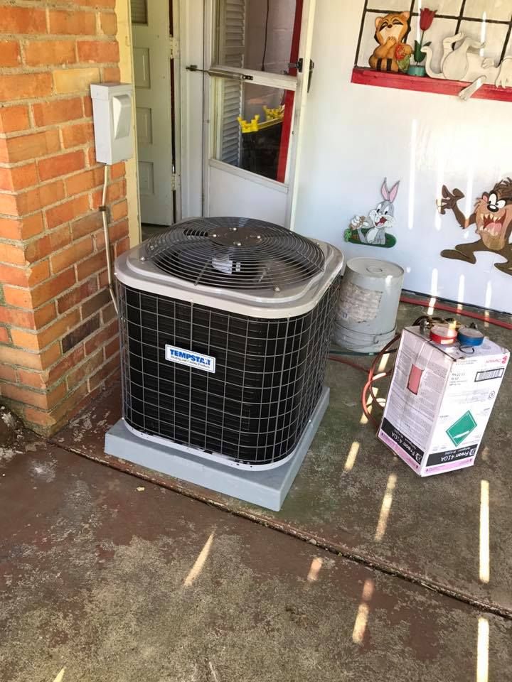 A Trane air conditioning unit sits on a concrete patio next to a brick wall, with a refrigerant tank beside it.