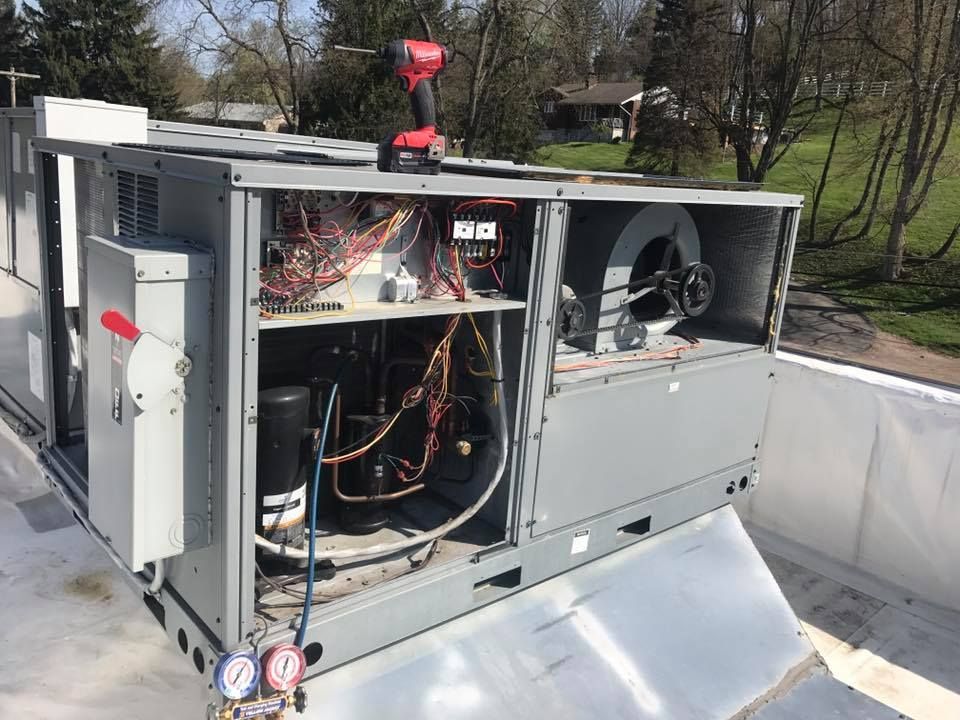 An open HVAC rooftop unit being serviced with a drill and manifold gauges visible on a sunny day.