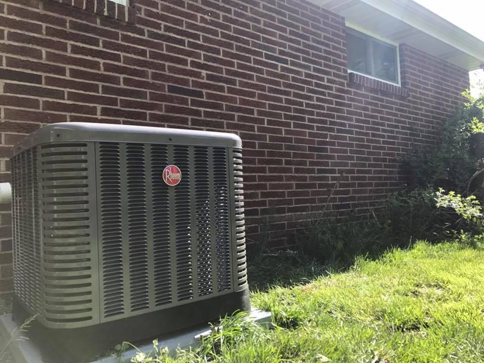 A grey Rheem outdoor air conditioning unit sits on a concrete pad against a brick house wall in a grassy yard.