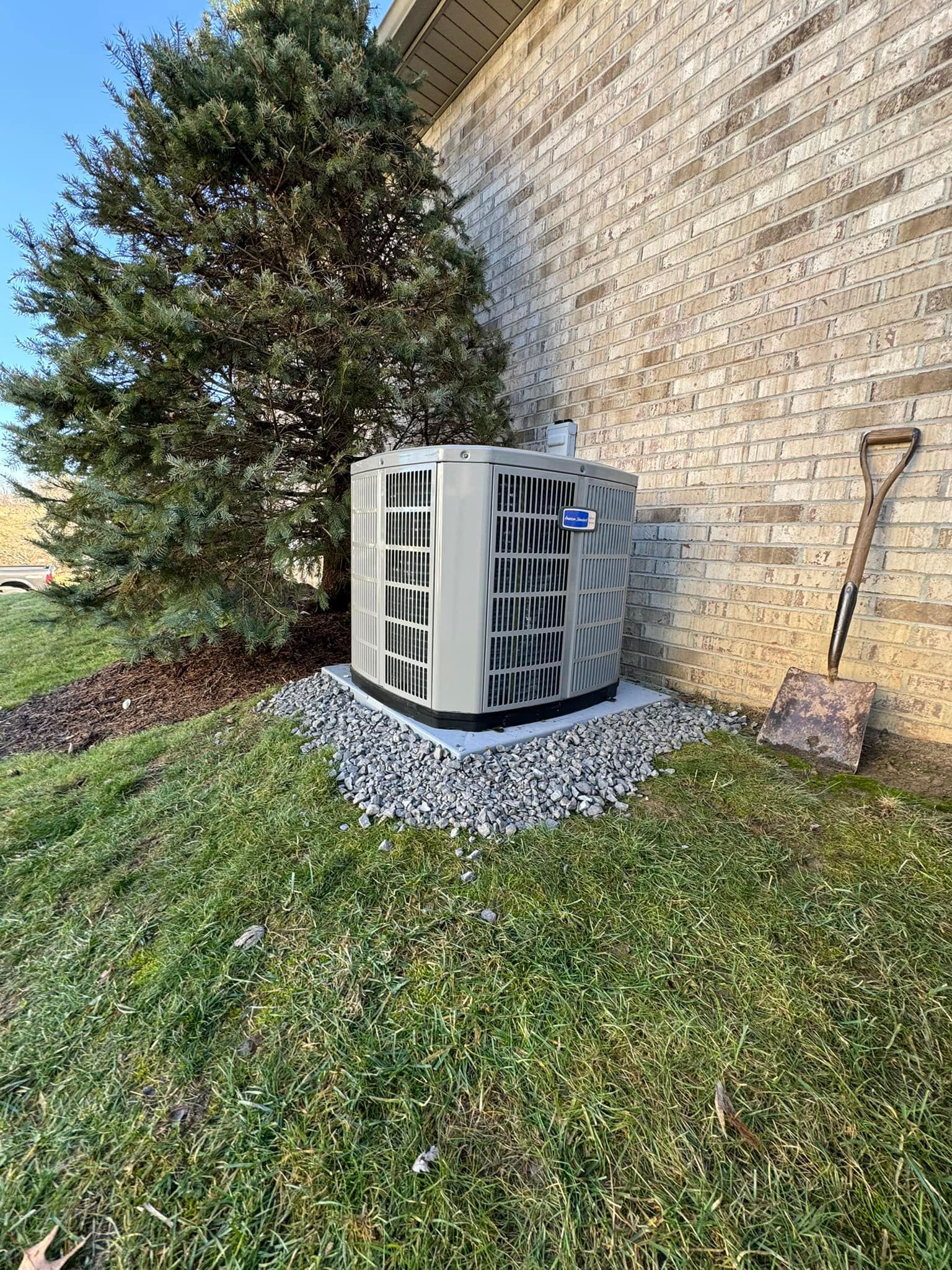 An outdoor HVAC unit sitting on a gravel pad next to a brick house and an evergreen tree, with a shovel leaning nearby.