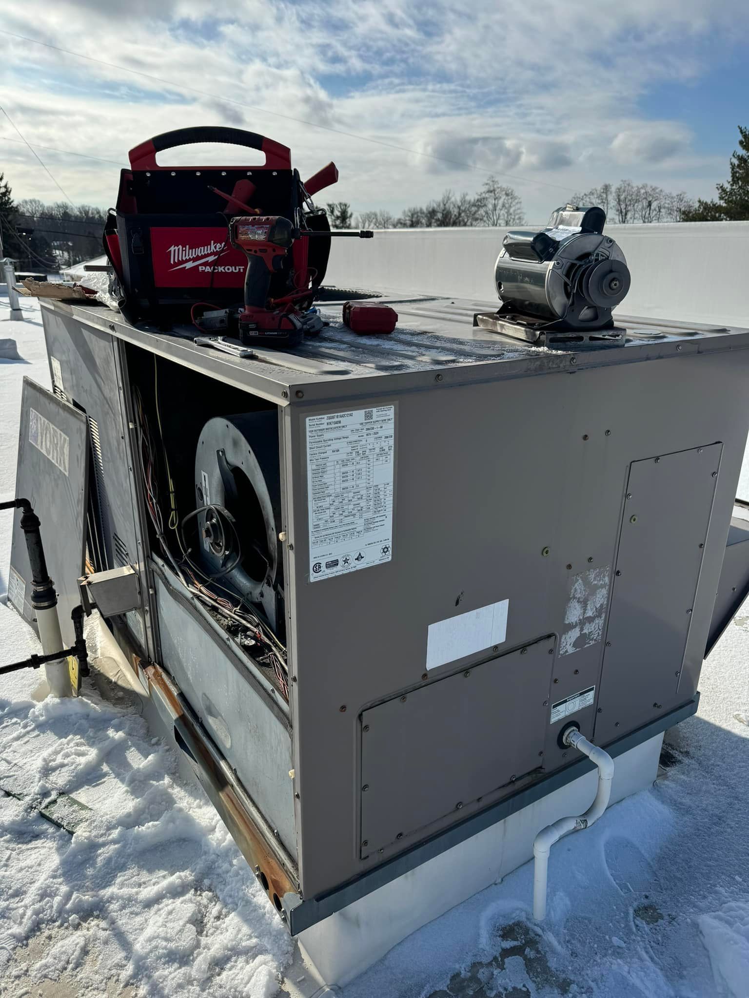 A technician's tool bag and a new blower motor sitting on a rooftop HVAC unit during winter, surrounded by snow.