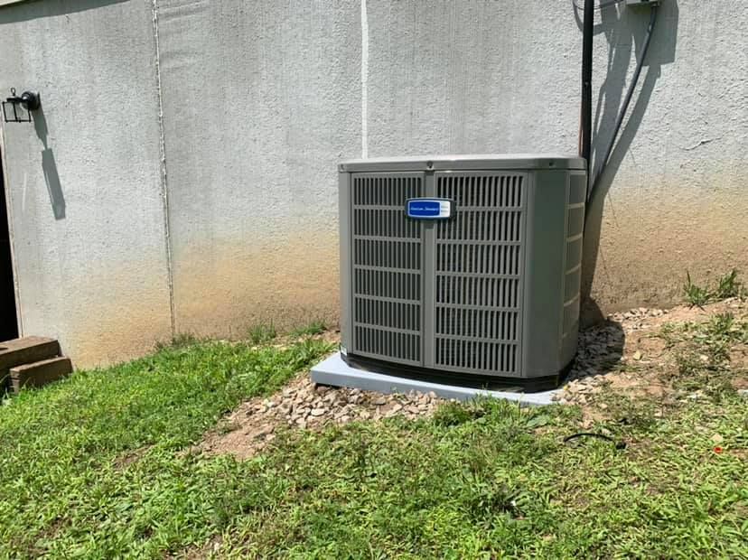 A gray rectangular outdoor air conditioning unit sits on a concrete pad against a plain, light-colored exterior wall.