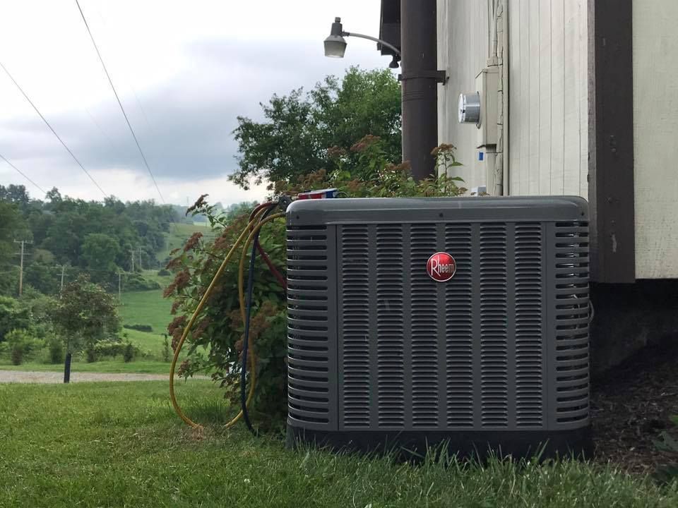 A gray Rheem air conditioning unit sits on a lawn next to the side of a house, with service gauges attached.