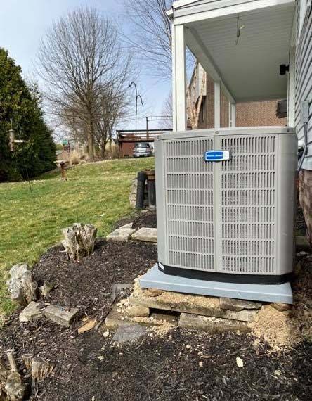 An outdoor central air conditioning unit sits on a concrete pad near a house porch in a yard with trees and mulch.