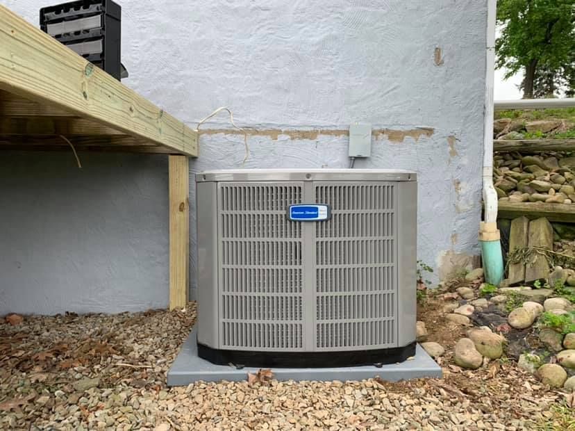 An outdoor gray air conditioning unit sits on a gravel base next to a house wall under a wooden deck.