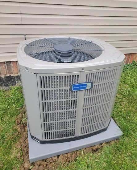 A beige American Standard air conditioning unit sits on a concrete pad against a house with light-colored siding.