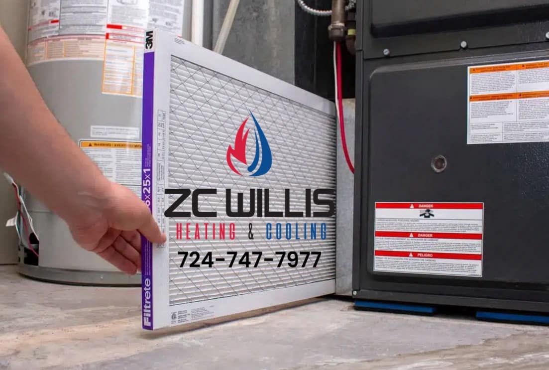 A person holds a large white HVAC air filter for ZC Willis Heating & Cooling next to a home furnace.
