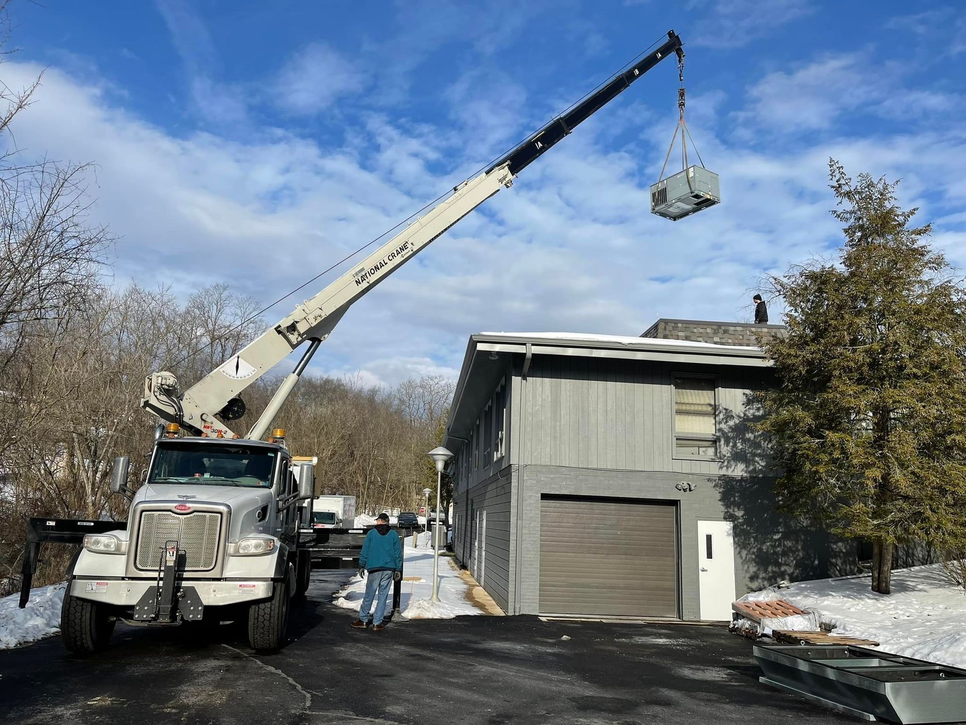 A crane lifts an HVAC unit onto the roof of a two-story gray building on a sunny, snowy day.