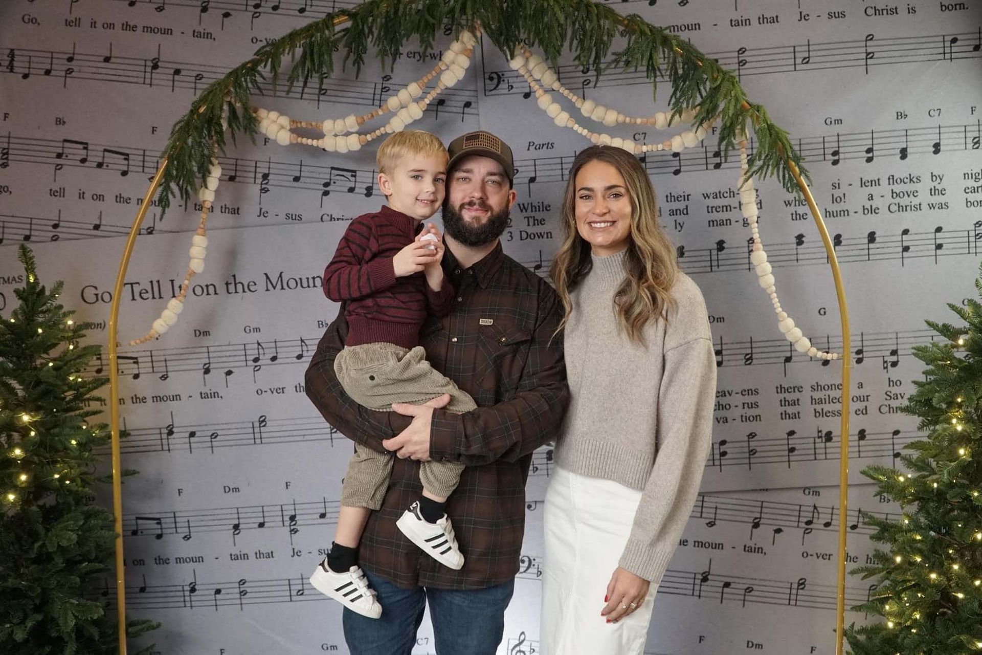 A family of three poses together under an arched garland, backed by a sheet music backdrop and flanked by Christmas trees.