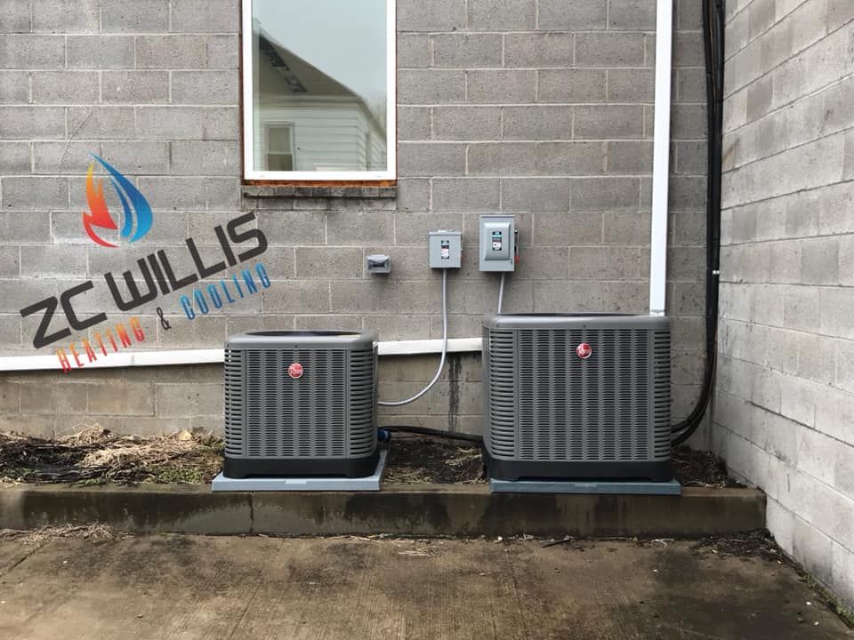 Two grey AC units sit on concrete pads against a brick wall with a logo for ZC Willis Heating & Cooling on the left.